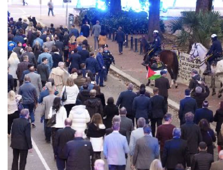 Wide view of a crowd marching down a street past mounted police, one protester carrying a Palestinian flag.